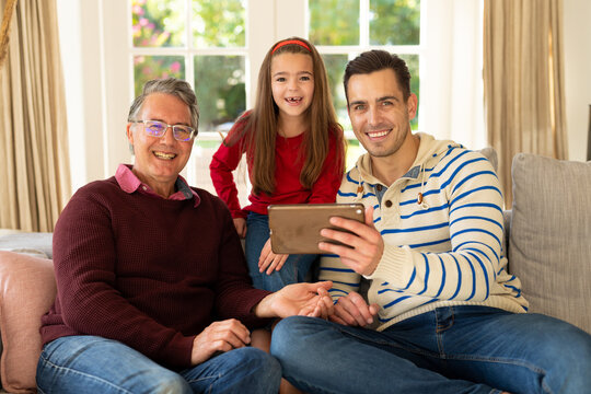Image of happy caucasian grandfather, father and daughter sitting on sofa with tablet