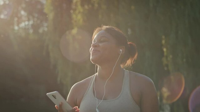 Plus sized African-American woman walking and using mobile phone with earphones in the park in a summer day 
