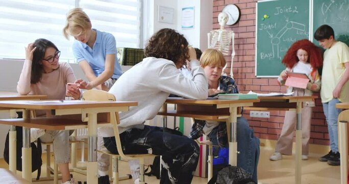 Caucasian teenagers, scholboys and schoolgirls sitting at desks and talking while having break at school from lessons. Teens boys and girls chatting and playing while resting in classroom.