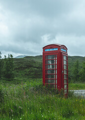 British vintage red phone booth in the middle of nowhere, along a country road in Scotland. overcast cloudy day, rain drizzle. Contrast between technology and nature, red and green.