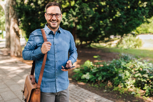 Portrait of smiling male executive walking on his way to work. Young professional commuter with smart phone is carrying laptop bag. He is wearing formals in the the city park.