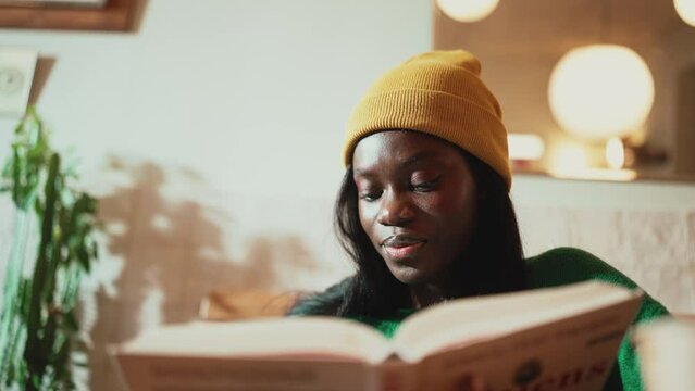 Smiling African young woman wearing yellow hat reading book and looking at the camera in cafe