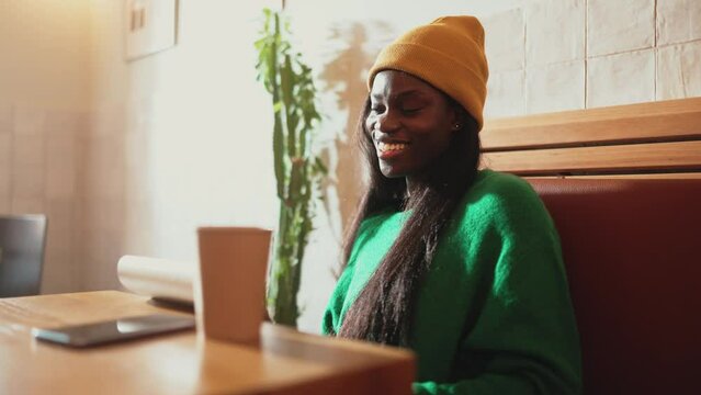 Laughing African young woman wearing yellow hat writing something in notepad while sitting in cafe