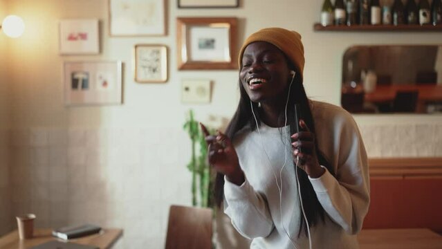 Smiling African young woman wearing white sweater singing and dancing in cafe