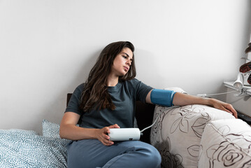 Young sick woman or girl measures her blood pressure with a digital blood pressure monitor. Ill female lying in the bed high blood pressure. - obrazy, fototapety, plakaty