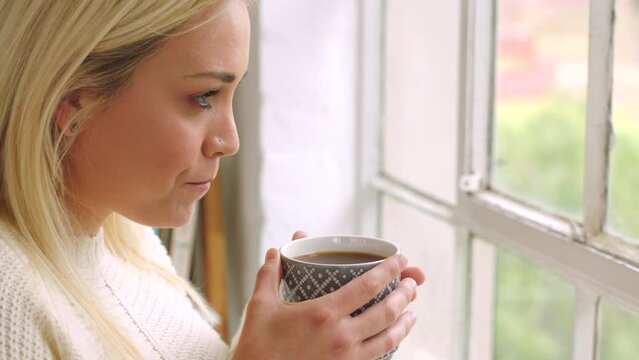 A beautiful young woman enjoying a warm cup of coffee and smiling in the morning. One happy blonde female drinking tea, looking at the view from a window in her apartment and thinking about the day