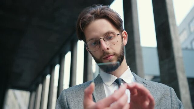 Close up of Caucasian bearded man wearing glasses, suit and tie using futuristic smartphone with transparent screen. Concept of future. Hologram. Touching device