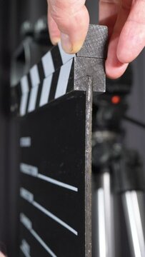 Vertical video social media format – Closeup of a man’s hands using a traditional wooden clapperboard in front of a studio video camera on a tripod, ready to shoot an inside scene take.