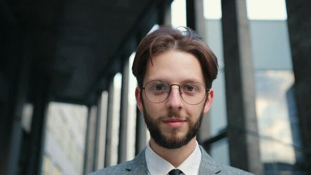 Close up of Caucasian brunette man putting on glasses, looking and smiling at camera. Portrait of successful cheerful bank worker, employee wearing suit. Concept of success.