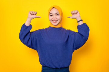 Cheerful young Asian Muslim woman dressed in casual sweater pointing at herself with proud isolated over yellow background