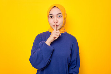 Serious young Asian Muslim woman dressed in casual sweater showing shh gesture, asks to be quiet isolated over yellow background