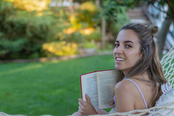 Young woman from the back looking to camera reading a book as leisure activity in the park of the city in spring