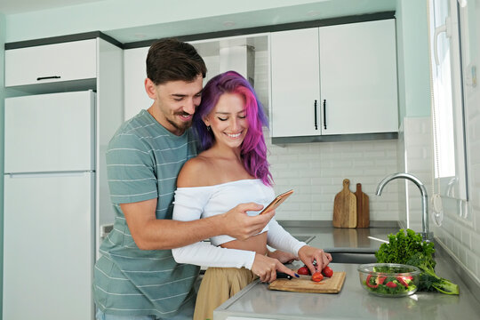Young woman with colorful hair and her husband cooking food in the open plan kitchen of their new home. Hipster couple spending quality time together. Copy space, interior background.