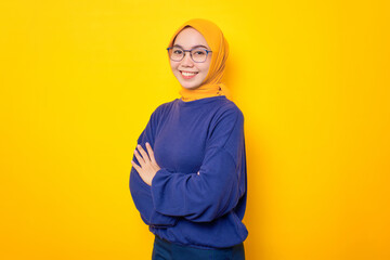 Fototapeta premium Smiling young Asian Muslim woman dressed in casual sweater crossed arms and looking confident isolated over yellow background