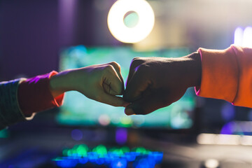 Indoor shot in a gaming environment in a dark place of two multiracial people making a fist bump in front of a monitor during an online game competition. High quality photo