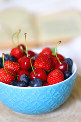 Bowl of strawberries, cherries and blueberries, open book and reading glasses on the bed. Selective focus.