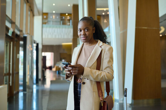 Young African American Woman In Smart Casualwear Looking At Camera While Standing In Hotel Lounge And Texting In Mobile Phone