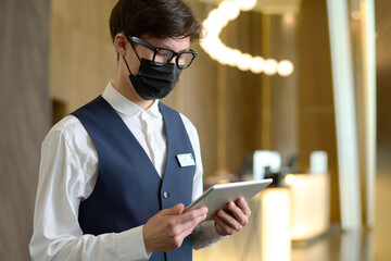 Young receptionist of modern hotel in uniform, eyeglasses and protective mask using tablet while...
