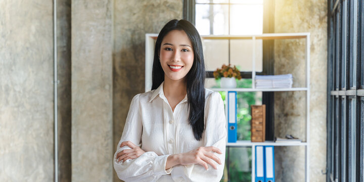 Portrait of smiling Asian millennial businesswoman stand in modern office hallway posing, happy confident young female employee or boss look at camera make picture in business workspace