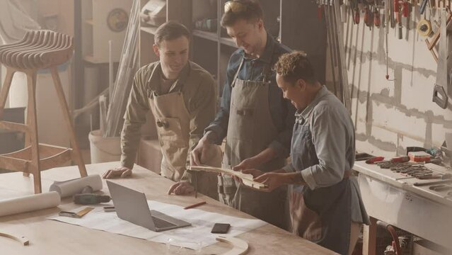 Team of carpenters in aprons looking at drawing plan on table while making wooden furniture in workshop