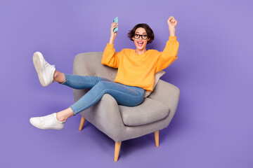 Portrait of attractive cheerful girl sitting using device having fun rejoicing isolated over violet lilac color background