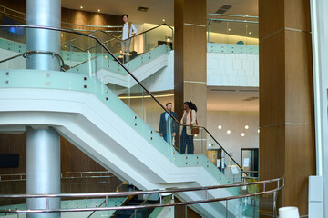 Two young multicultural colleagues in formalwear having talk while moving upstairs on staircase in hotel lounge during business travel