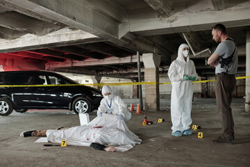 Young policeman communicating with forensic expert at crime scene with dead body covered with white...