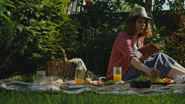 Young woman having summer picnic. Girl eating outside, reading book. Summer outdoors activities. Backyard picnic.