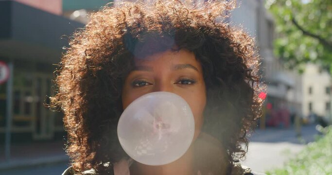 Young trendy girl blowing bubblegum outdoors. Cool, youthful Afro American woman with playful expression on face making a big bubble with chewing gum. Funky student having fun outdoors in a city park