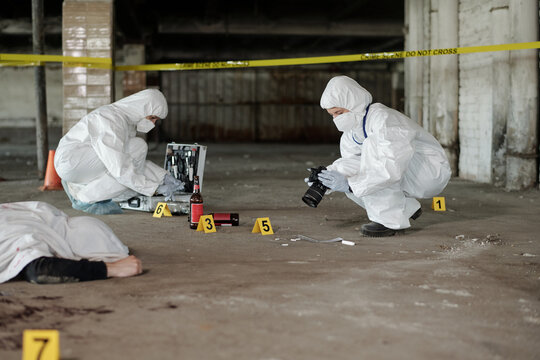 Two criminological experts in coveralls taking photos of evidences and inspecting crime scene surrounded by yellow tape in parking area