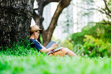Asian man in casual clothes reading under a tree, sitting on the grass and relaxing with a book in...