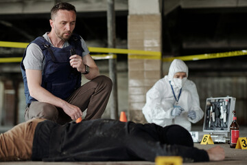 Young serious policeman in bulletproof vest squatting by dead body while inspecting crime scene...