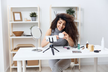 Professional makeup artist tanned handsome curly Latin beauty blogger in casual top sit at the table in home interior. Copy space Mockup Banner. Influencer record video review blog using smartphone