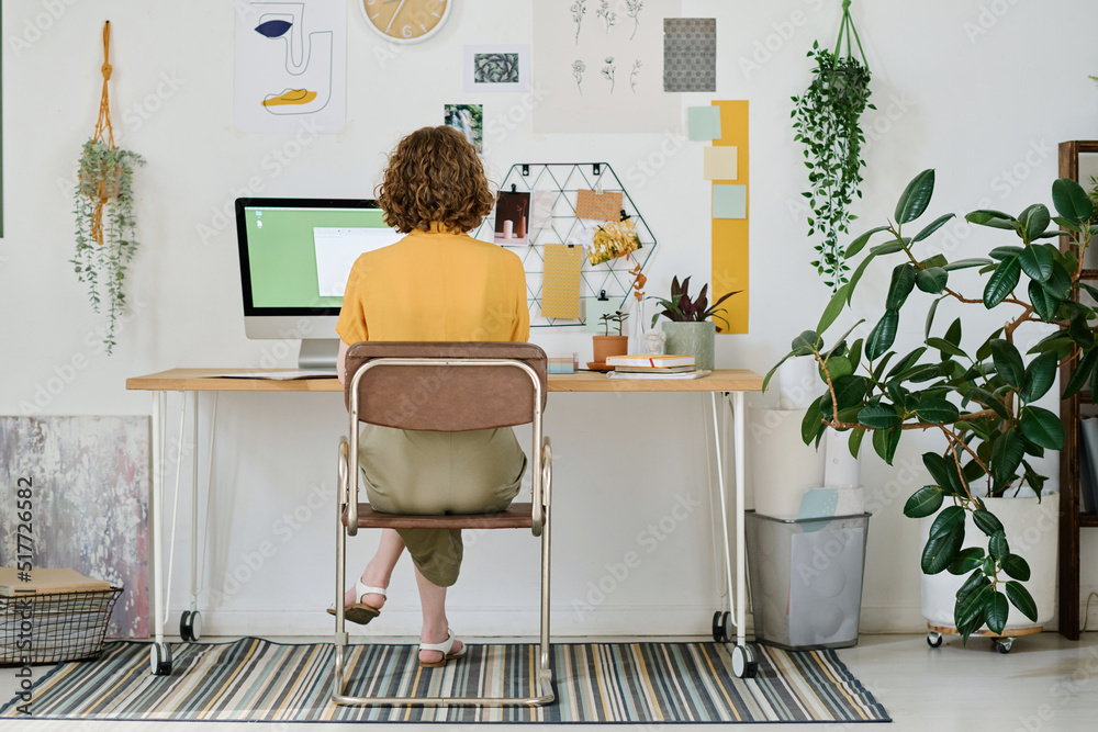 Poster Back view of young businesswoman or freelancer working in front of computer monitor while sitting by desk and creating new website