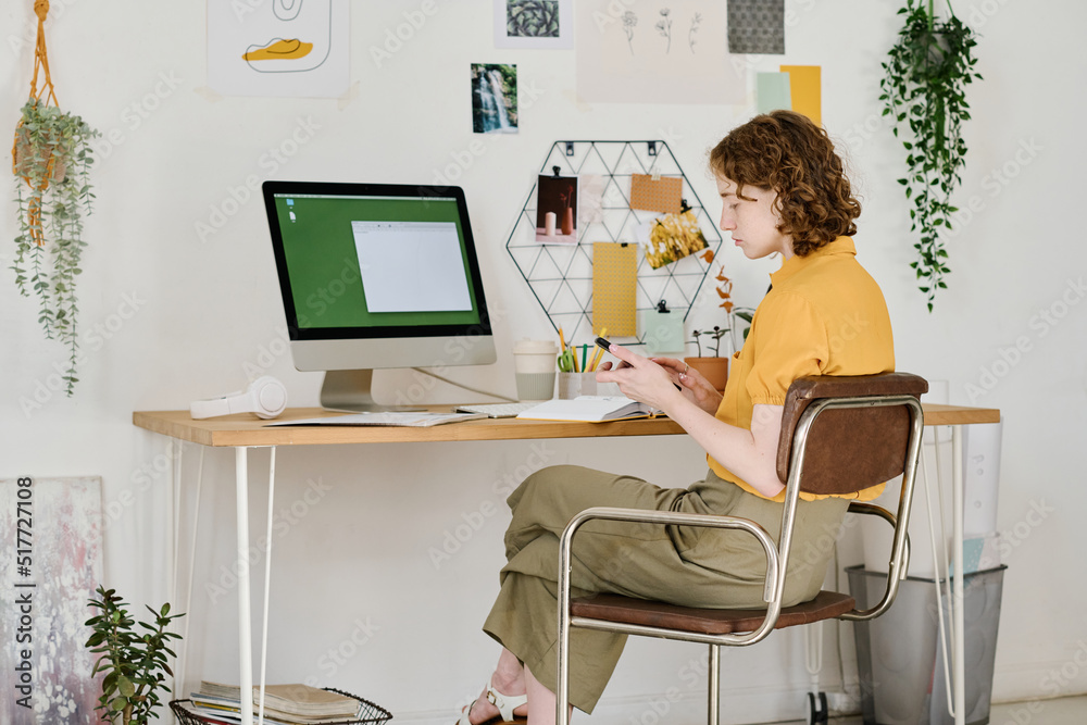 Poster young serious businesswoman scrolling in mobile phone while sitting in front of computer monitor and