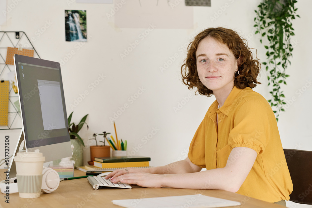 Poster young successful freelancer in yellow blouse looking at camera while sitting by workplace in front o