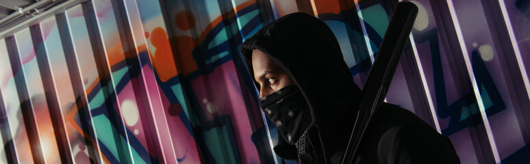 Side view of african american bandit in mask holding baseball bat near graffiti on wall, banner - obrazy, fototapety, plakaty