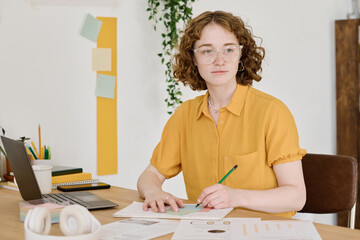 Young creative woman in eyeglasses and yellow blouse drawing schemes on paper while sitting by...