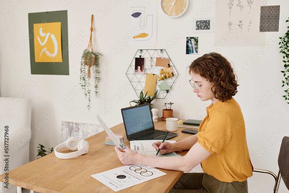 Poster young female accountant or economist making notes in notebook or on paper while looking through and 