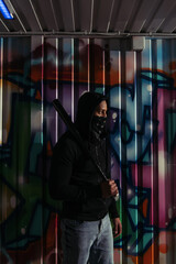 Fototapeta na wymiar Side view of african american hooligan in mask and hoodie holding baseball bat near graffiti on wall