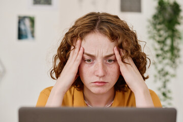 Young female freelancer with tense facial expression looking at laptop screen while sitting by...