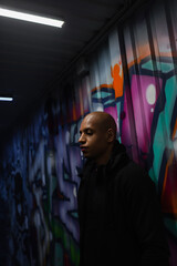 African american hooligan standing near graffiti in garage - obrazy, fototapety, plakaty