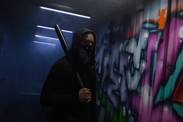 African american hooligan in mask holding baseball bat near graffiti and smoke in garage