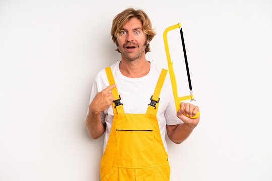 Adult Blond Man Looking Shocked And Surprised With Mouth Wide Open, Pointing To Self. Handyman With A Saw Concept