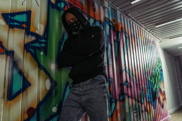 African american vandal in mask and hoodie crossing arms near graffiti on wall