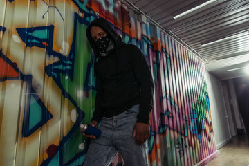 African american vandal in hood holding spray paint near graffiti on wall