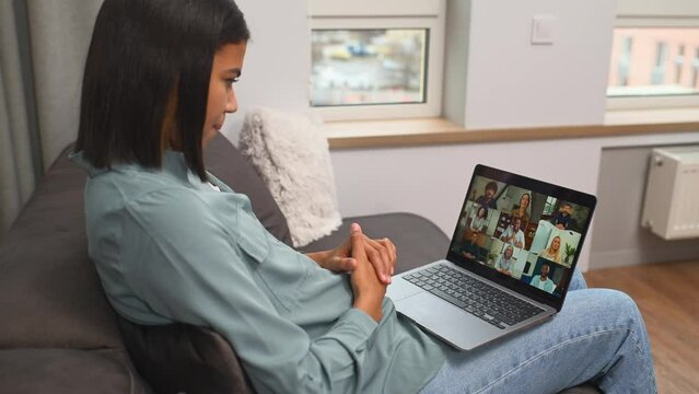 Remote meeting via video call. Asian woman sitting on the sofa and using app on laptop for online communication with group of diverse multiracial people. Side view