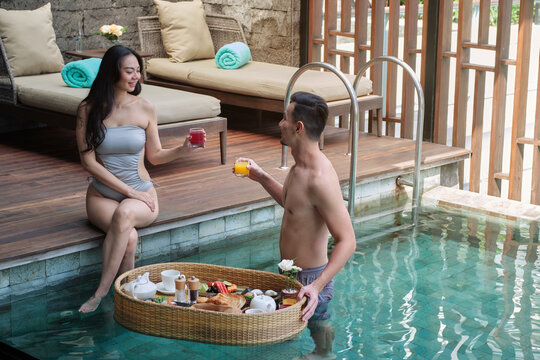 Happy Asian couple having breakfast in the pool. Floating breakfast tray in luxury hotel. Man and woman are drinking fresh juice and talking