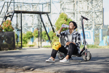 Asian man drinking water while sitting on electric scooter at the street. Modern and ecological transportation concept.