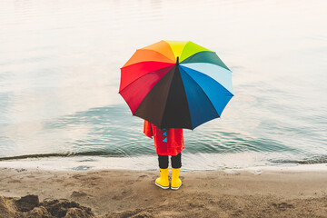 Boy in a red raincoat and yellow rubber boots holds rainbow umbrella standing at beach. Child with colourful umbrella looking at sea. School kid in a waterproof red coat holding multicolor umbrella.